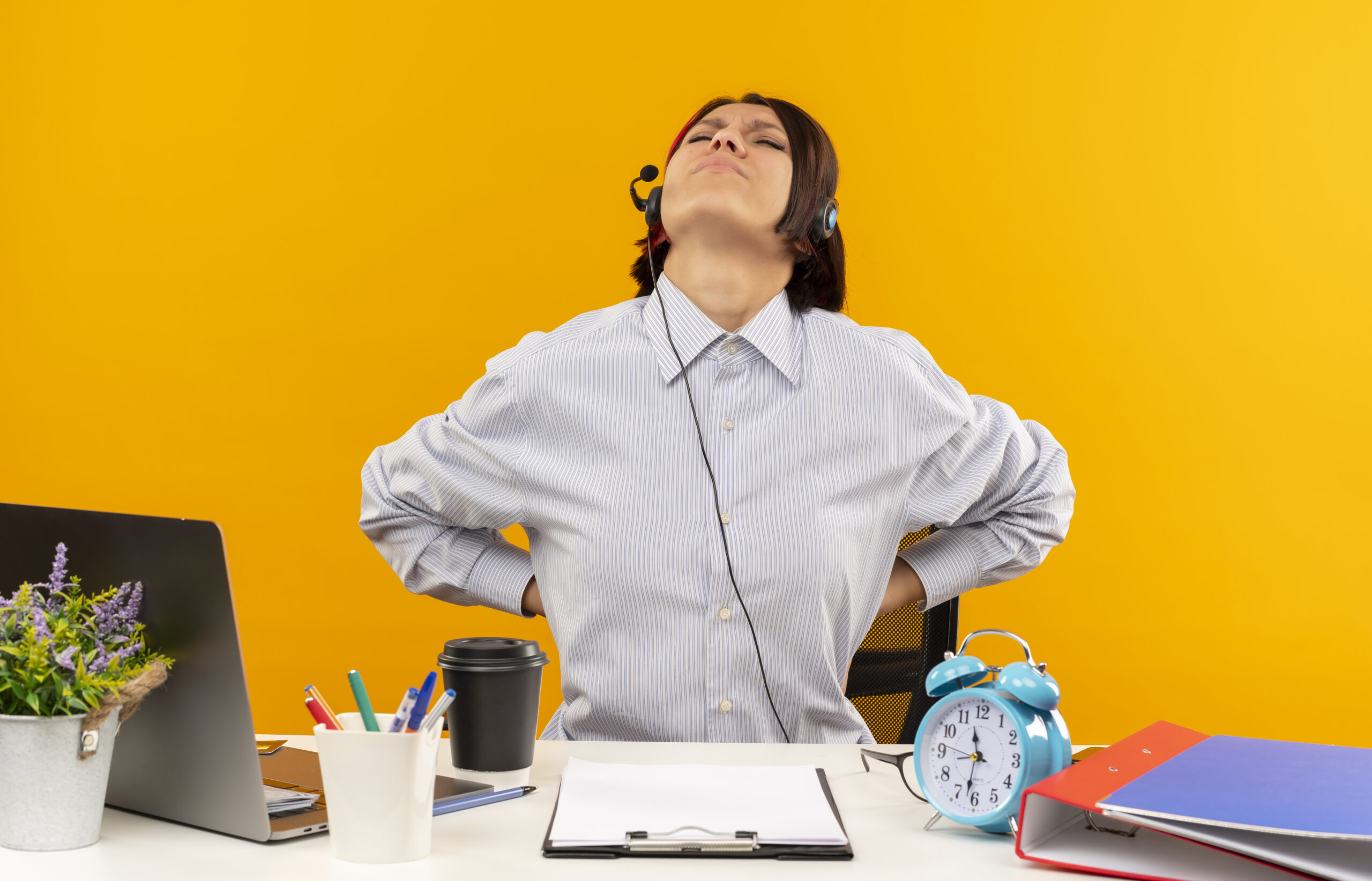 aching young call center girl wearing headset sitting at desk putting hands on back with closed eyes isolated on orange background