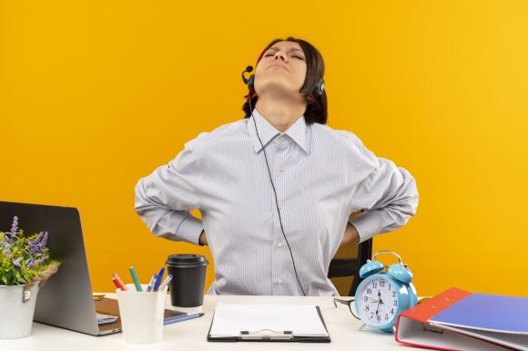 aching young call center girl wearing headset sitting at desk putting hands on back with closed eyes isolated on orange background